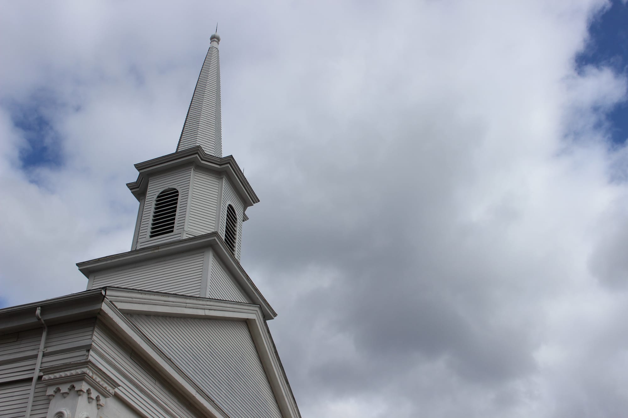 Steeple of wooden white country church with clouds surrounding it.